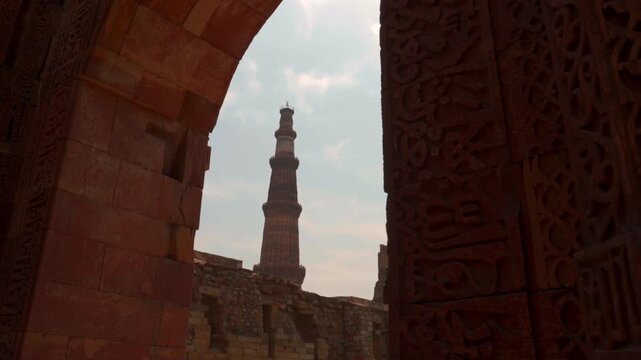 A panning shot revealing the Qutb Minar victory tower framed by an ancient red sandstone arch with intricate Islamic calligraphy and ruins in the background.