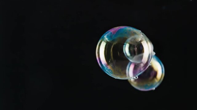 Numerous soap bubbles with vibrant iridescent colors drift and combine against a solid black backdrop. The light reflects off their surfaces, creating a dynamic and abstract visual effect.
