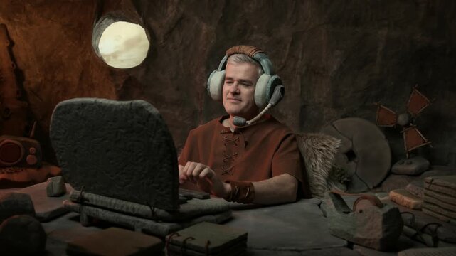 Confident prehistoric man working in his office, he is wearing a headset, answering calls and typing on the laptop: business and customer assistance in the stone age concept