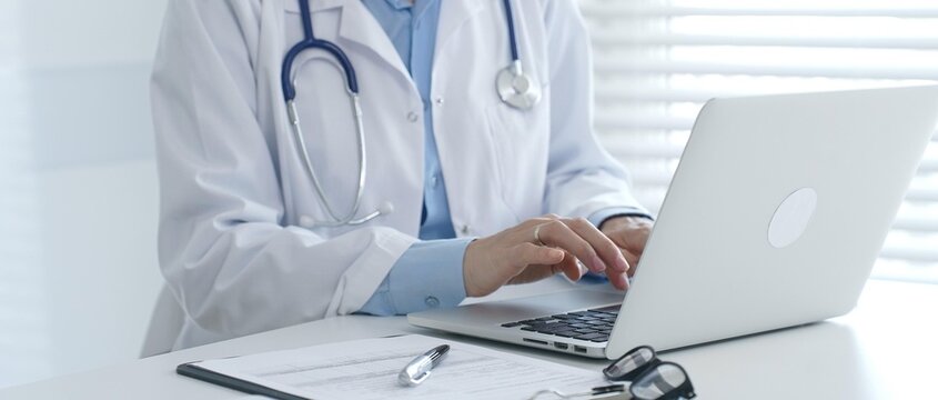 Female doctor documents patient information while working with digital devices at minimalist white workstation in medical office. Medicine and healthcare concept