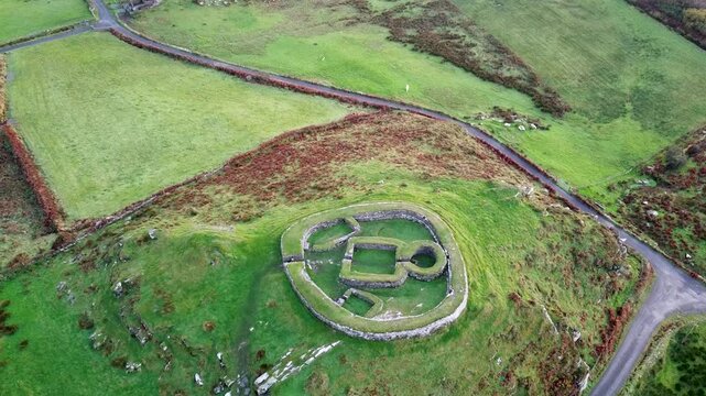 Aerial shot descending down on the Leacanabuaile Ringfort, the ancient historic fort with square internal ruins, surrounded by green grass and autumn brush