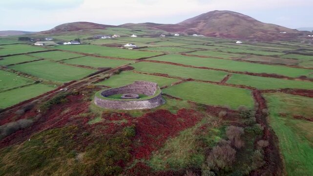 Aerial drone shot orbiting the circular stone walls of the ancient Cahergal Ringfort, vibrant autumn landscape background of green pastures and distant mountains in Kerry, Ireland