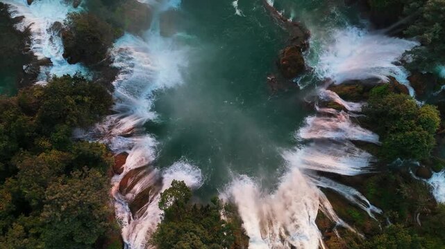 Cinematic drone shot ascending and turning over the majestic Ban Gioc Waterfall. Cascading white water falls into a turquoise river surrounded by lush tropical forest in Northern Vietnam.