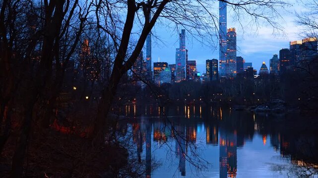 Night view of Manhattan skyline reflecting in Central Park lake. City lights and skyscraper windows reflecting in dark water through silhouette of bare trees at night or dusk.
