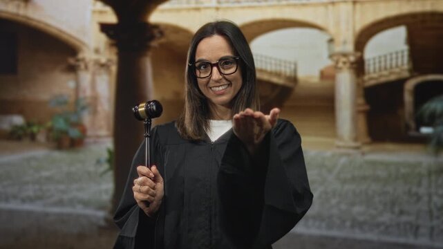 Woman judge in black robe holds gavel and offers open hand gesture toward viewer in a historic court building, smiling with glasses; confidence.