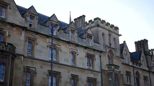 Building at Oxford University with traditional college facade, dormer windows, and stone walls along a city street, classic Oxford architecture captured in wide urban view