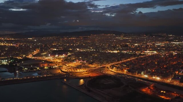 Breathtaking aerial footage of the illuminated athens cityscape at night, showcasing the vibrant city lights, the port of piraeus, and the peace and friendship stadium under a cloudy sky