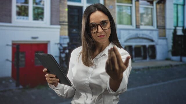 Woman in white shirt wearing glasses holding a tablet extends hand palm up in beckoning gesture on a street with building facade; confidence invitation.