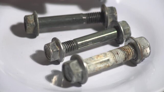 Detailed 4K shot of several long metallic engine bolts with nuts being placed on a white surface. These used fasteners show signs of wear, suitable for automotive assembly, construction, mechanical re