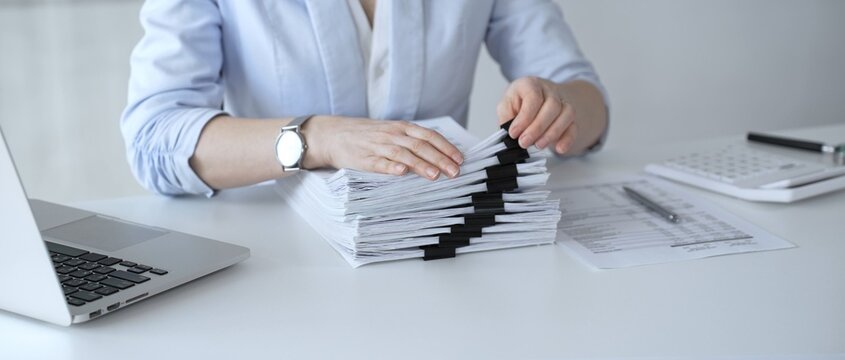 Business person working on financial documents, accounting or auditing a large stack of paperwork, managing tax forms and corporate reports with a pen at an office desk. Audit and taxes