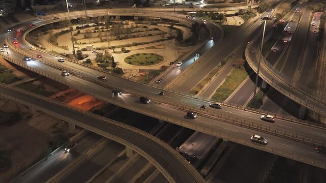 Aerial view of a multilevel highway interchange at night, with illuminated roads and cars with headlights on creating light trails as they navigate the complex network of overpasses and ramps