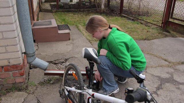 Diligent young girl in casual clothing crouching in the backyard and carefully inflating a flat bicycle tire with a hand pump, preparing for a ride on a cloudy day outdoors