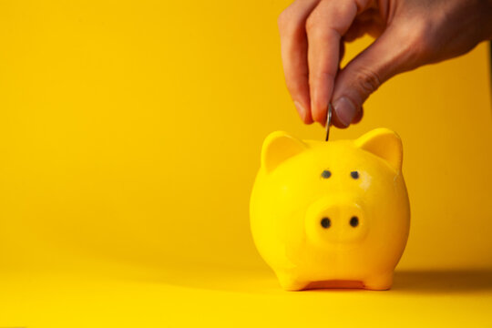Female hand putting coin into a yellow piggy bank on a yellow background