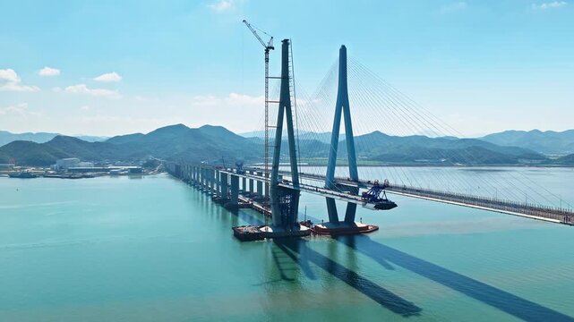 Aerial shot of a large cable-stayed bridge under construction with heavy machinery over the blue sea in Xiangshan, Ningbo, China