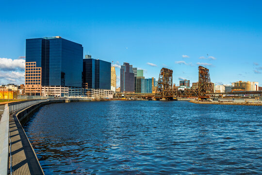 A vibrant skyline of Newark, New Jersey, featuring modern glass office buildings and the historic Dock Bridge reflecting over the Passaic River under a clear blue sky.