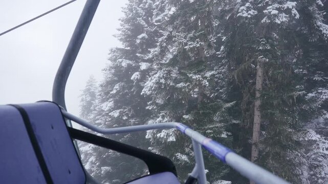 POV from ski lift moving through snowfall above snowy pine forest, winter resort atmosphere, alpine mountains, cold foggy weather, scenic winter travel