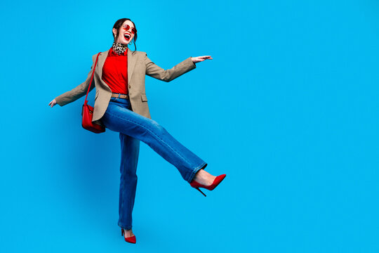 Joyful fashion woman in red heels and blazer posing against vibrant blue background, stylish casual outfit with handbag and sunglasses expressing confidence and playful movement