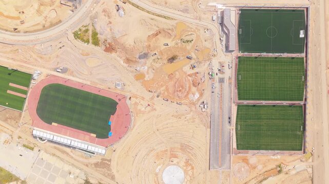 High-angle drone perspective showing the development of a large sports complex, including a track and field stadium, soccer fields, and an outdoor amphitheater under construction