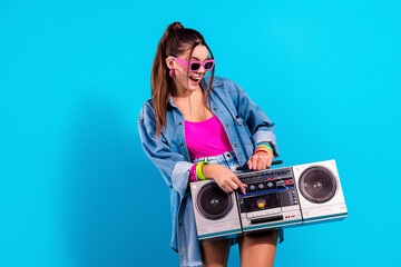 Young woman with boombox in retro 80s style outfit holding cassette player while wearing pink sunglasses and denim jacket, vibrant pop fashion portrait conveying fun and energetic music vibes © deagreez