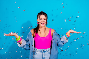 Fototapeta premium Young woman celebrating with confetti in pink top and denim jacket, joyful party portrait on blue background