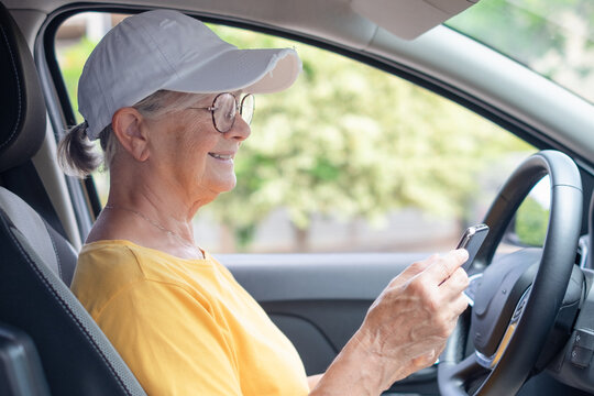 Senior woman stops driving the car to type a message on her smartphone, elderly female satisfied with journey by car, sits on driver`s seat. People, driving, transport concept
