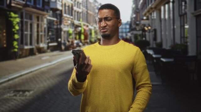 Young black man in yellow sweater holding smartphone and squinting with one hand raised in a puzzled shrug on a narrow street; frustration.