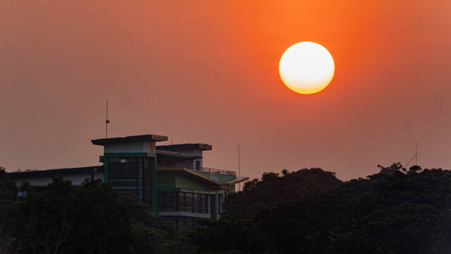 Bright sun over orange sunset sky above modern villa and treetops, silhouetted architecture at dusk with warm light, scenic peaceful landscape and copy space