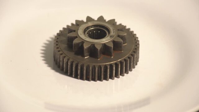 Close up of a metallic double spur gear wheel rotating slowly on a white isolated surface, showing detailed industrial machinery part.
