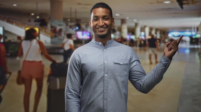 Black man pointing finger at arrivals board with visible hand and button shirt in airport; confidence welcome.