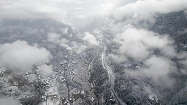 Drone flying over snowy mountain village with river in gorge among clouds, Svaneti, Georgia. Winter landscape, Caucasus mountains, aerial scenic view, misty atmosphere