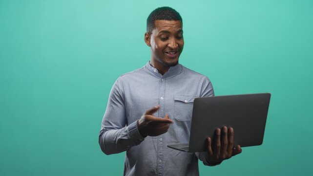 Man holding a laptop in one hand and pointing at the screen with the other, wearing a buttoned shirt against a teal studio backdrop; confidence communication.