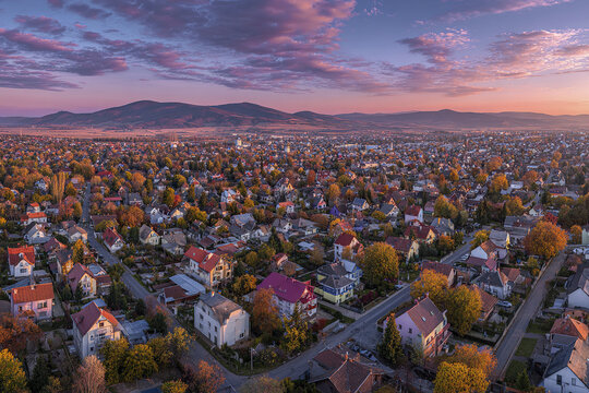 Small city aerial view with residential houses and autumn trees under colorful sunset sky, surrounded by distant mountains creating peaceful and scenic atmosphere