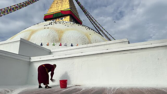 Slow motion of a Buddhist monk sweeping near Boudhanath Stupa in Kathmandu, Nepal. Early morning sunlight, prayer flags and flying birds create a peaceful spiritual atmosphere