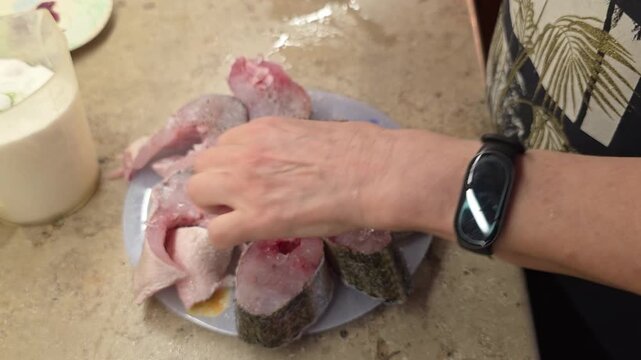 Woman seasoning fresh raw fish pieces with salt and pepper on plate in kitchen. Home cooking preparation of fish. Hand adding spices to seafood before cooking.