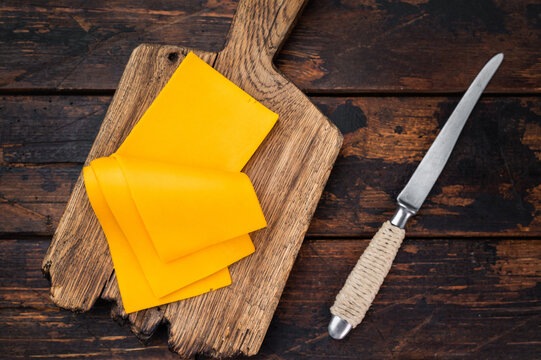Sliced cheddar style cheese on wooden cutting board with knife over rustic table background and copy space for food design