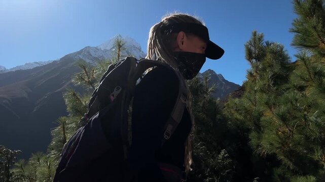 Female trekker with backpack records selfie video while climbing steep trail in Himalayan mountains, Nepal, then turns to camera, snowy peaks, Everest Base Camp route, high altitude adventure