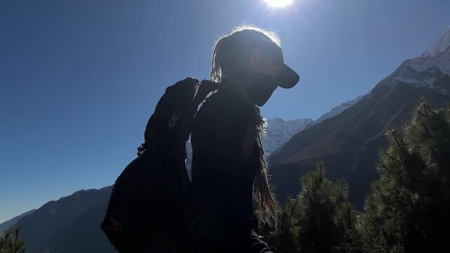 Female trekker with backpack records selfie video while climbing steep trail in Himalayan mountains, Nepal, then turns to camera, snowy peaks, Everest Base Camp route, high altitude adventure