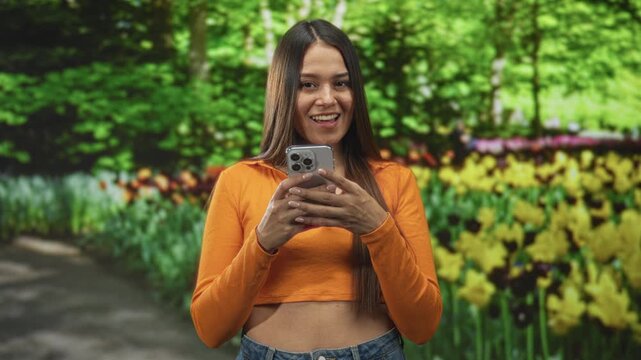 Woman holding smartphone, smiling and typing with midriff visible in a flowered forest path; joy connection.