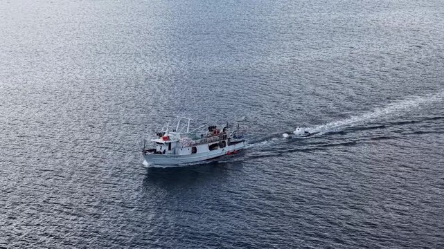 Aerial shot of a working fishing boat moving through dark blue water with a small boat behind it.