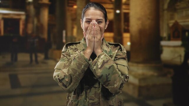 Woman soldier in camouflage uniform covers mouth with hands inside a cathedral building, framed by columns and an altar; relief gratitude.