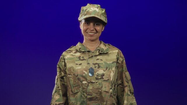 Woman soldier in camouflage uniform smiles and gives thumbs up, dog tags visible, slight head turn in studio; pride duty.