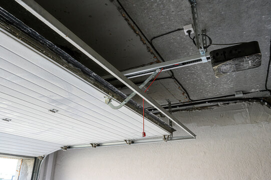 sectional garage door with a mounted opener mechanism; metal tracks fixed to the ceiling, red emergency cord visible; white door panels, electrical wiring connected to the motor unit
