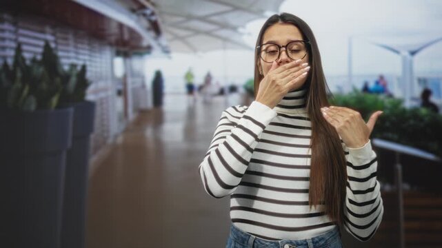 Woman covering mouth with hand, glasses visible, wearing striped turtleneck and jeans in an airport terminal near large planters and a canopy walkway; surprise secrecy.