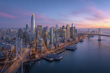 Fototapeta premium San Francisco city skyline panorama with Bay Bridge and waterfront at sunset, featuring modern skyscrapers and calm water under colorful sky