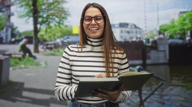 Woman holds book and pen and sticks tongue out while writing on a street canal walkway, wearing glasses and striped turtleneck; playful curiosity.