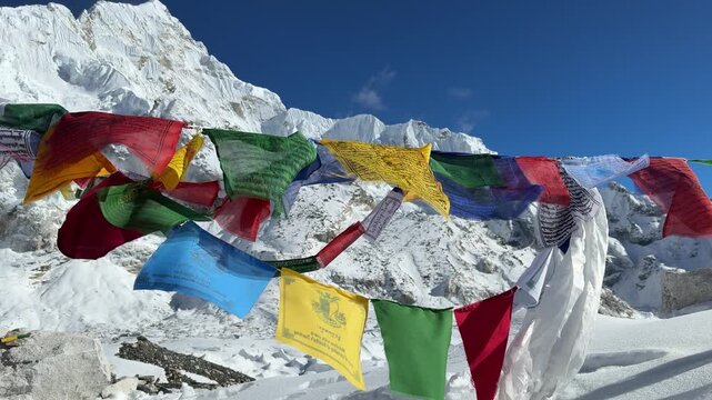 Prayer flags waving in the wind with Mount Everest in the background near Everest Base Camp, Nepal. Snow-covered Himalayan peaks under blue sky, spiritual atmosphere, travel, slow motion