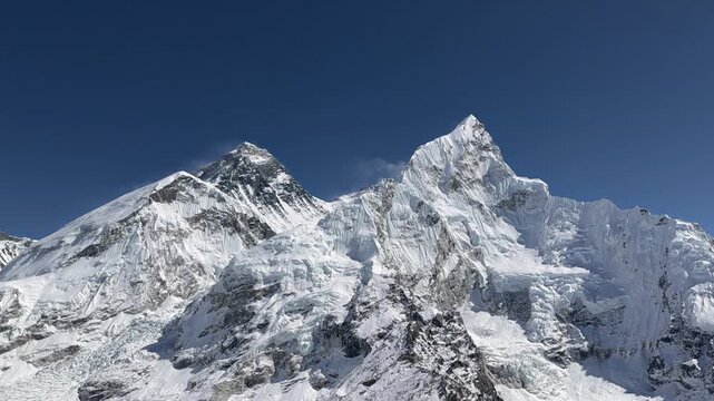 Mount Everest and surrounding Himalayan peaks under clear blue sky with moving clouds. Static shot showing detailed mountain relief and valley landscape in bright daylight, Nepal