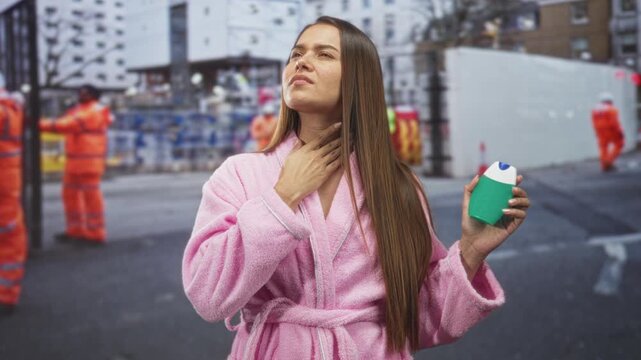 Woman in pink bathrobe holding a green toothpaste bottle and touching her throat on a street with construction workers; discomfort morning routine.
