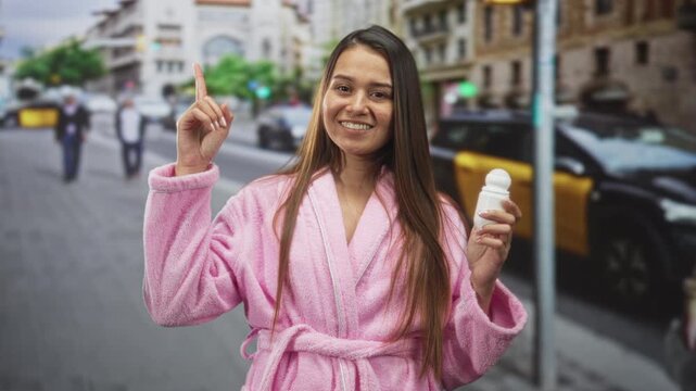 Young brunette woman in pink bathrobe holding roll on deodorant in her right hand and points index finger upward on street while smiling; confidence selfcare.
