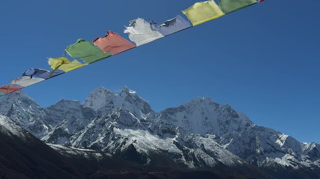 Prayer flags waving in the wind above Himalayan mountain peaks. Snow-covered mountains under blue sky, peaceful spiritual atmosphere, nature, travel, slow motion